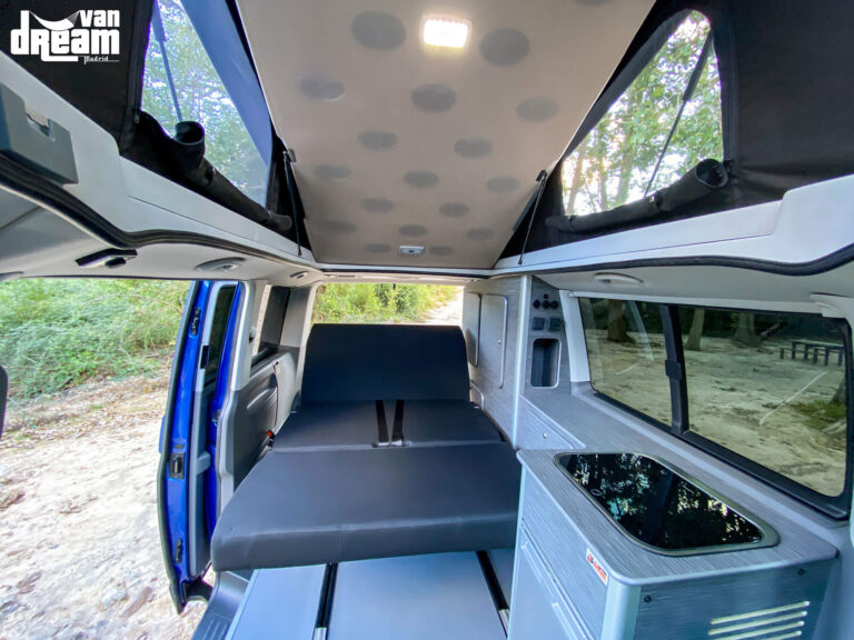 Vista interior de una Volkswagen T6.1 Caravelle con techo elevable BRAM abierto y cama superior elevada, mostrando la amplitud y altura disponible en el habitáculo.