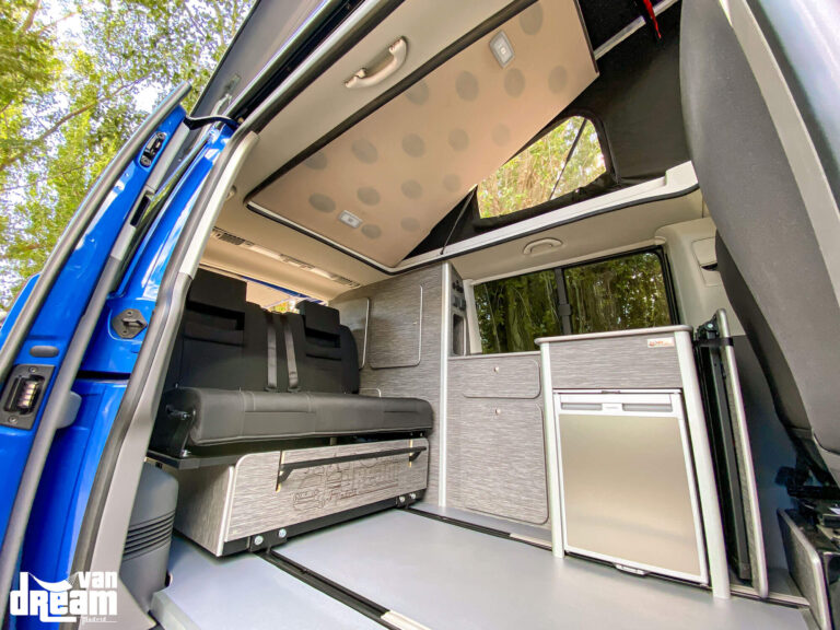 Vista lateral del interior de una Volkswagen T6.1 Caravelle con techo elevable BRAM y cama superior elevados, mostrando la amplitud del habitáculo y el mobiliario camper personalizado.