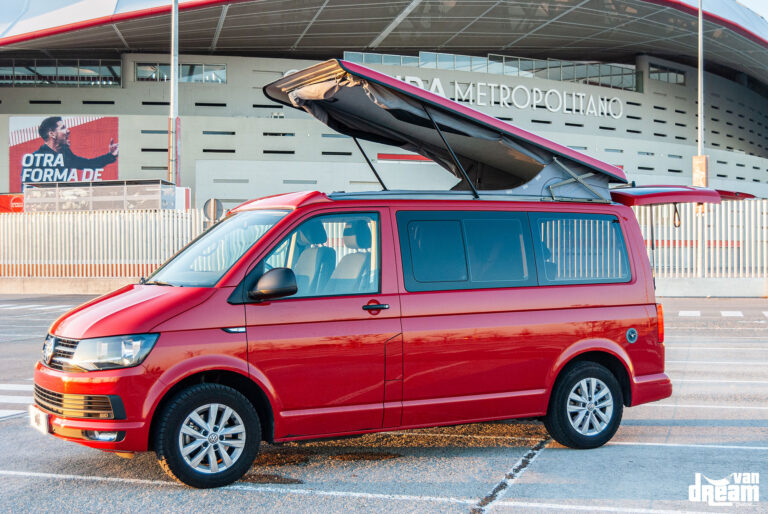 Vista lateral de una Volkswagen T6 Multivan roja con techo elevable Reimo abierto, lona gris recogida y estructura de elevación visible.